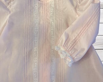Könnte beinhalten: Ein blassrosa Kleid mit langen Ärmeln und zwei vertikalen weißen Spitzenbändern auf der Vorderseite. Das Kleidungsstück verfügt über Biesen, einen Rundhalsausschnitt und weiße Spitzenbesätze an den Ärmelbündchen. Mit Knopfverschluss am Rücken.