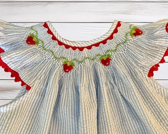 Könnte beinhalten: Ein hellblau-weißes Seersucker-Kleid mit gesmokter Passe, bestickt mit roten Erdbeeren und grünen Ranken. Das Kleidungsstück hat Flatterärmel mit rotem Wellensaum und einen runden Ausschnitt.