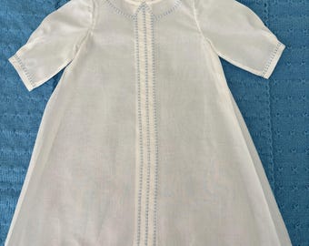 Könnte beinhalten: Ein weißes Kleid mit kurzen Ärmeln und einem runden Kragen. Das Kleidungsstück ist mit vertikalen Reihen hellblauer Ziernähte entlang der vorderen Mitte, am Kragen, an den Ärmelbündchen und am unteren Saum verziert.