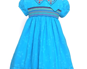 Könnte beinhalten: Ein leuchtend blaues Kleid mit einem dezenten, tonalen Blumenmuster, kurzen Puffärmeln und einem Bubikragen mit bunter Blumenstickerei. Das Oberteil ist mit horizontalen Reihen aus mehrfarbigen Smok-Details versehen.
