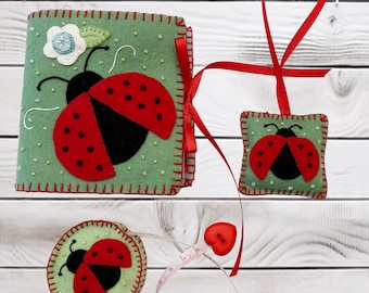 Könnte beinhalten: Ein Set aus drei Filzobjekten mit Marienkäfer-Applikationen: ein quadratisches Nadelbuch mit weißer Blume, ein kleiner quadratischer Anhänger und ein rundes Nadelkissen. Alle Teile sind salbeigrün mit roten Zierstichen und rotem Band.