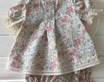 Könnte beinhalten: Ein zweiteiliges Set bestehend aus einer kurzärmeligen Bluse und passender Pumphose. Der Stoff zeigt ein Blumenmuster in Rosa, Blau und Grün auf cremefarbenem Grund. Die Bluse hat drei Knöpfe, Puffärmel mit Spitzenbesatz und Bändern sowie einen gesäumten Spitzenrand. Die Pumphose hat elastische Beinabschlüsse mit Spitzenbesatz.