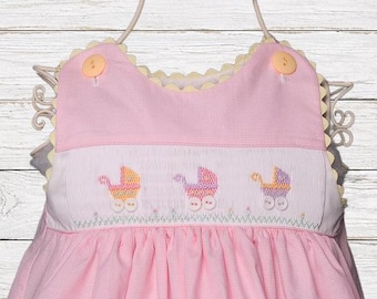Könnte beinhalten: Ein rosa Kleid mit Vichy-Muster und einem weißen, gesmokten Brustbereich, der mit gestickten Kinderwagen in Gelb, Rosa und Violett verziert ist. Das Kleid hat einen gewellten Ausschnitt, zwei pfirsichfarbene Knöpfe an den Schultern und weiße Knöpfe an den Kinderwagen.