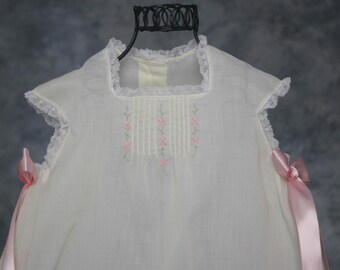 Könnte beinhalten: Ein weißes ärmelloses Kleid mit quadratischem Ausschnitt und Spitzenbesatz, vertikalen Biesen auf der Brust und kleiner rosa Blumenstickerei. Die Seiten sind mit langen, hellrosa Satinbändern verziert, die zu Schleifen gebunden sind.