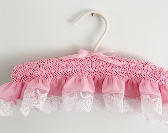 Könnte beinhalten: Ein gepolsterter Kleiderbügel, der mit rosa Stoff bezogen ist und oben ein aufwendiges Smok-Detail aufweist. Der Bügel hat eine rosa Satinschleife in der Mitte und einen gerüschten rosa Stoffbesatz am unteren Rand, verziert mit weißer Spitze.