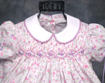Könnte beinhalten: Ein weißes Kleid mit rosa-violettem Blumenmuster, einem einfarbigen weißen Bubikragen, kurzen Puffärmeln mit lavendelfarbenen Schleifen und aufwendigen violetten Smok-Details auf der Brust mit gestickten Blumenakzenten.