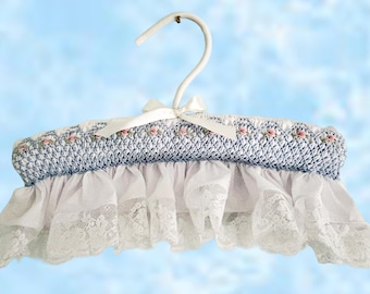 Könnte beinhalten: Ein gepolsterter Kleiderbügel, bezogen mit blauem Stoff in Smock-Optik und kleiner rosa Blumenstickerei. Der Bügel ist oben mit einer weißen Satinschleife verziert und hat am unteren Rand einen weißen Rüschenbesatz mit Spitzenkante.