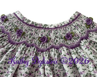 Könnte beinhalten: Ein weißes Kleid mit kleinem violettem Blumenmuster, einem gesmokten Einsatz mit violettem Zickzack-Stickmuster und kleinen violetten gehäkelten Rosenapplikationen mit grünen Blättern. Der Text Kathy Dykstra © 2026 ist violett aufgedruckt.