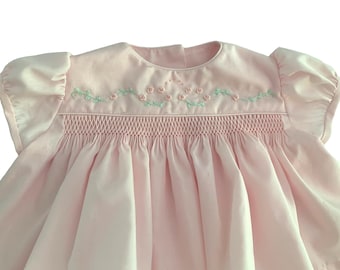 Könnte beinhalten: Ein hellrosa Kleid mit kurzen Puffärmeln und einem Rundhalsausschnitt. Das Oberteil zeigt ein komplexes Smok-Detail und eine zarte Stickerei aus kleinen rosa Rosen mit hellgrünen Blättern im Brustbereich.