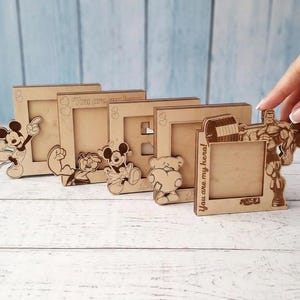 May include: A collection of wooden photo frames with laser-cut designs. The frames feature cartoon characters like Mickey Mouse and a weightlifter. Some frames have the text "You are my hero!" and "You are my love".