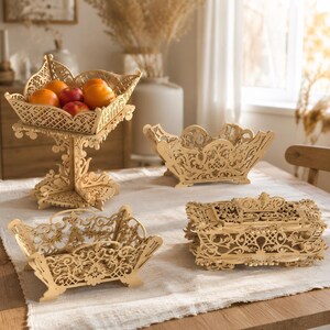 May include: A collection of four light-colored wooden items featuring intricate laser-cut patterns. The set includes a tall pedestal fruit bowl filled with oranges and apples, two open-work rectangular baskets, and a rectangular lidded box.