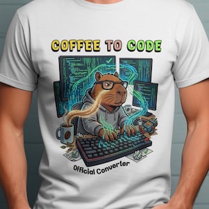 Può includere: T-shirt bianca con un'illustrazione a cartoni animati di un capibara con occhiali e una felpa con cappuccio, che digita su una tastiera. Il design include schermi di computer, caffè e il testo "COFFEE TO CODE" e "Official Converter."