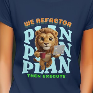 Op de afbeelding: Marineblauw T-shirt met een cartoon leeuw die een mok en een laptop vasthoudt. De tekst luidt "WE REFACTOR PLAN PLAN THEN EXECUTE" in oranje, wit en groen. De leeuw is bruin en bruin.