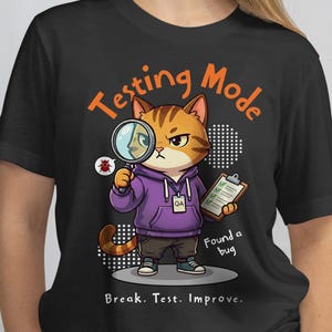 May include: Black t-shirt featuring a cartoon cat wearing a purple hoodie and holding a magnifying glass. The text "Testing Mode" and "Found a bug" are displayed. The phrase "Break. Test. Improve." is also present.
