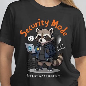 May include: Black t-shirt with a cartoon raccoon dressed as a security guard, holding a tablet displaying a lock icon. The text "Security Mode" and "Protect what matters" are in orange and white.