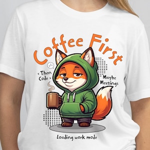 May include: White t-shirt featuring a cartoon fox in a green hoodie holding a coffee mug. The text "Coffee First" and "Loading work mode" are in orange. Additional text includes "Then Code" and "Maybe Meetings".