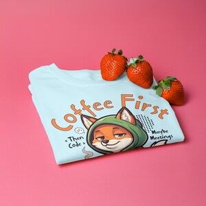 Könnte beinhalten: Hellblaues T-Shirt mit einem Cartoon-Fuchs mit grüner Kapuze. Der Text auf dem Shirt lautet "Coffee First, Then Code, Maybe Meetings". Drei rote Erdbeeren liegen auf dem Shirt.