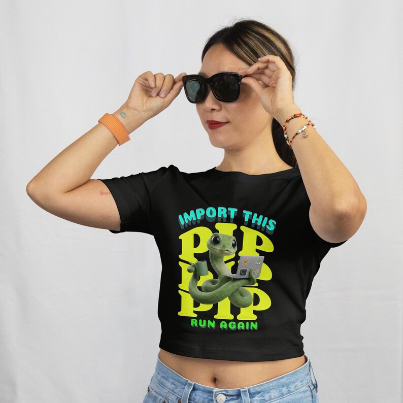 May include: Black crop top with the text "IMPORT THIS PIP RUN AGAIN" in teal and yellow. The shirt features a cartoon green snake holding a laptop. The model is wearing black sunglasses and blue jeans.
