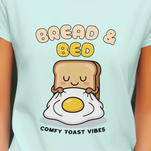 May include: Pale blue t-shirt with a cartoon design. The graphic shows a slice of toast embracing a fried egg, with the words "BREAD & BED" above and "COMFY TOAST VIBES" below. The toast and egg have smiling faces.