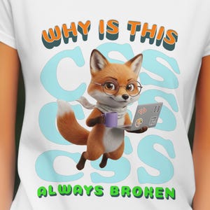 May include: White t-shirt featuring a cartoon fox wearing glasses and a scarf, holding a laptop and a purple mug. The shirt has the text "WHY IS THIS CSS ALWAYS BROKEN" in orange, blue, and green.