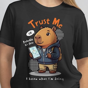 May include: Black t-shirt featuring a cartoon capybara wearing a hoodie and holding a tablet and pen. The text "Trust Me" and "I know what I'm doing" are displayed in orange and white. A speech bubble says "ok Probably it's fine".