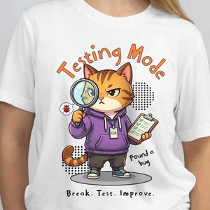 May include: White t-shirt featuring a cartoon cat wearing a purple hoodie and holding a magnifying glass. The cat is inspecting a bug, with the words "Testing Mode" and "Found a bug" printed on the shirt. The text "Break. Test. Improve." is also printed on the shirt.