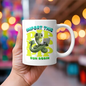 May include: White ceramic mug with a green snake design, holding a laptop and a coffee cup. The mug features the text "IMPORT THIS PIP PIP PIP RUN AGAIN" in green and blue. The mug has a white handle.