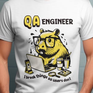 Può includere: T-shirt bianco con un castoro giallo dei cartoni animati con gli occhiali e che lavora su un laptop. Il testo "QA ENGINEER" e "I break things so users don't" sono stampati sulla maglietta. Una tazza di caffè, un telefono e una lente d'ingrandimento sono anche nell'immagine.