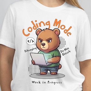 May include: White t-shirt featuring a cartoon bear with a laptop. The bear is in "Coding Mode" with text that says "Debugging" and "Do Not Disturb". The phrase "Work in Progress" is also displayed.