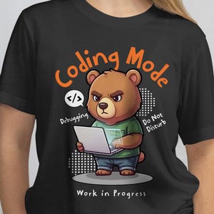 May include: Black t-shirt featuring a cartoon bear with a laptop. The bear is wearing a green shirt and blue jeans. Text on the shirt reads "Coding Mode", "Debugging", "Do Not Disturb", and "Work in Progress".