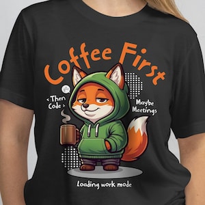 May include: Black t-shirt featuring a cartoon fox wearing a green hoodie and holding a coffee mug. The shirt has the text "Coffee First" and "Loading work mode". Additional text includes "Then Code" and "Maybe Meetings".