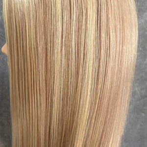 May include: A mannequin head with long, straight, blonde hair. The hair has subtle highlights and lowlights, creating a natural, multi-dimensional look. The hair is styled with a center part and falls to the shoulders.
