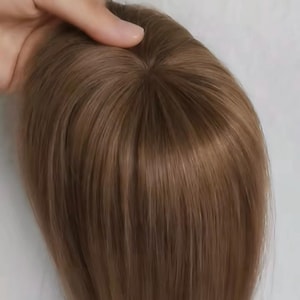 Pode incluir: Uma touca de cabelo castanho claro com mechas longas e lisas. A peça de cabelo foi projetada para adicionar volume e cobertura. O cabelo é de um tom quente e natural, e a base é visível. A peça de cabelo é segurada por uma mão.