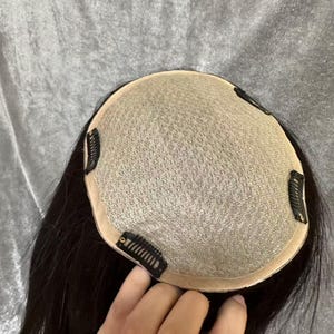 May include: A black hair topper with a mesh base and several attached clips. The hair is dark and appears to be straight. The topper is designed to add volume or coverage to the existing hair.