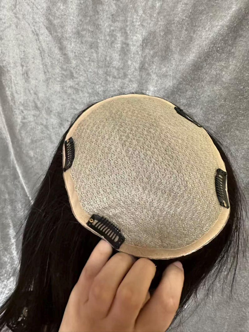 May include: A black hair topper with a mesh base and several attached clips. The hair is dark and appears to be straight. The topper is designed to add volume or coverage to the existing hair.