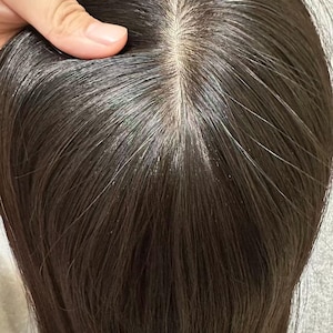 Op de afbeelding: Een donkerbruine haartop met een natuurlijk ogende scheiding. Het haar is steil en lijkt van synthetische vezels te zijn gemaakt. De topper wordt door een hand omhoog gehouden, die de bovenkant van het hoofd en de textuur van het haar laat zien.