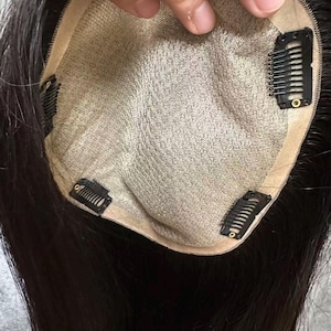 May include: A dark brown hair topper with a beige mesh base and several black clips for attachment. The hair is long and straight, with a natural appearance. The topper is designed to add volume and coverage.