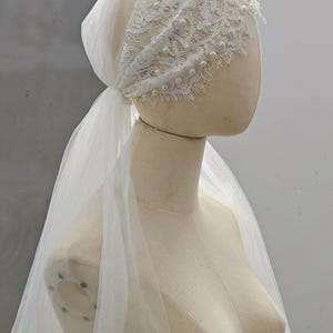 Puede incluir: Un velo de novia blanco y un tocado en un maniquí. El tocado presenta un diseño de encaje adornado con perlas. El velo está hecho de una tela transparente y fluida. El texto "Jennie Veil Co." es visible.