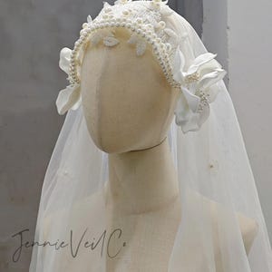 Puede incluir: Un velo de novia con un tocado decorativo. El tocado presenta un diseño de perlas y encaje con flores de tela. El velo es transparente y adornado con pequeños detalles de perlas. El texto "Jennie Veil Co." es visible.