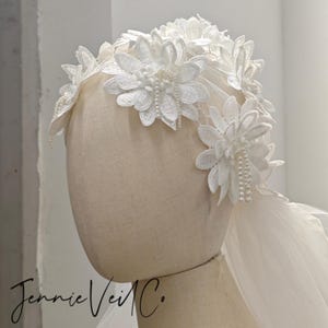 Puede incluir: Tocado de novia blanco con grandes y delicadas flores de encaje con detalles de perlas. Las flores están dispuestas en la parte superior de la cabeza, con un velo transparente en la parte posterior. El tocado se muestra en una cabeza de maniquí.