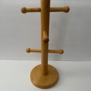 May include: A light brown wooden mug tree stand featuring a central vertical post attached to a circular base. The stand includes six horizontal pegs with rounded ends arranged in pairs at different heights to hold mugs or other hanging items.