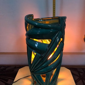 May include: A teal ceramic table lamp with a cut-out leaf pattern that emits a warm yellow glow from within. A yellow measuring tape is visible behind the lamp, showing a height measurement reaching approximately 45 centimeters.