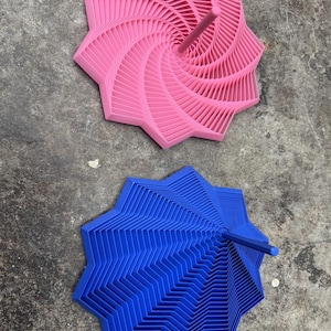 May include: Two abstract, star-shaped objects. One is bright pink, the other is blue. Both have a spiral or layered design with a central stem. The objects are on a gray, textured surface.