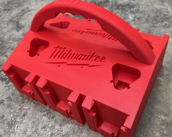 May include: A red battery caddy with a handle and the Milwaukee logo. The caddy has multiple slots and a rectangular shape. The text "BATTERY CADDY" is at the bottom of the image.