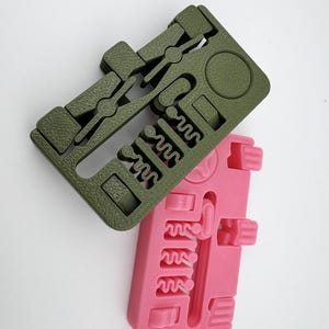 Könnte beinhalten: Zwei 3D-gedruckte Objekte in Olivgrün und Rosa. Das grüne Objekt hat eine rechteckige Form mit verschiedenen Aussparungen, darunter ein Kreis und ein Zickzackmuster. Das rosa Objekt hat ein ähnliches Design. Beide Objekte scheinen ineinandergreifende Teile zu sein.