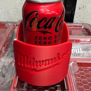 Puede incluir: Una lata roja de Coca-Cola Zero en un portavasos rojo de Milwaukee. La lata tiene el logo de Coca-Cola en negro y las palabras "ZERO SUGAR" en blanco. El portavasos tiene el logo de Milwaukee en blanco.