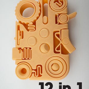 Op de afbeelding: Een perzik-kleurig 12-in-1 multi-tool met diverse vormen en uitsparingen, waaronder tandwielen, cirkels en een handvat. De tool heeft een getextureerd oppervlak, met de tekst "12-in-1" in zwart onderaan gedrukt.