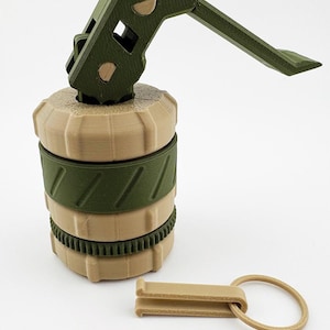 Op de afbeelding: Een 3D-geprinte granaatreplica in olijfgroen en beige. Het ontwerp omvat een hendel, een pin en een cilindrisch lichaam met geribbelde en gebandeerde secties. Er is ook een aparte beige pin met een ring.
