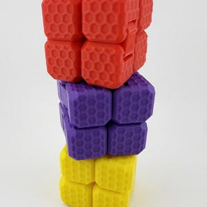 May include: A three-tiered fidget toy in red, purple, and yellow. Each tier is composed of four connected cubes with a honeycomb pattern. The toy is designed to be manipulated and played with, offering a tactile experience.