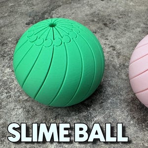 Op de afbeelding: Een groene, bolvormige slijmbal met verticale groeven en een decoratieve bovenkant. De tekst "SLIME BALL" staat in het wit, omlijnd met zwart, onderaan de afbeelding. De bal ligt op een grijs, getextureerd oppervlak.